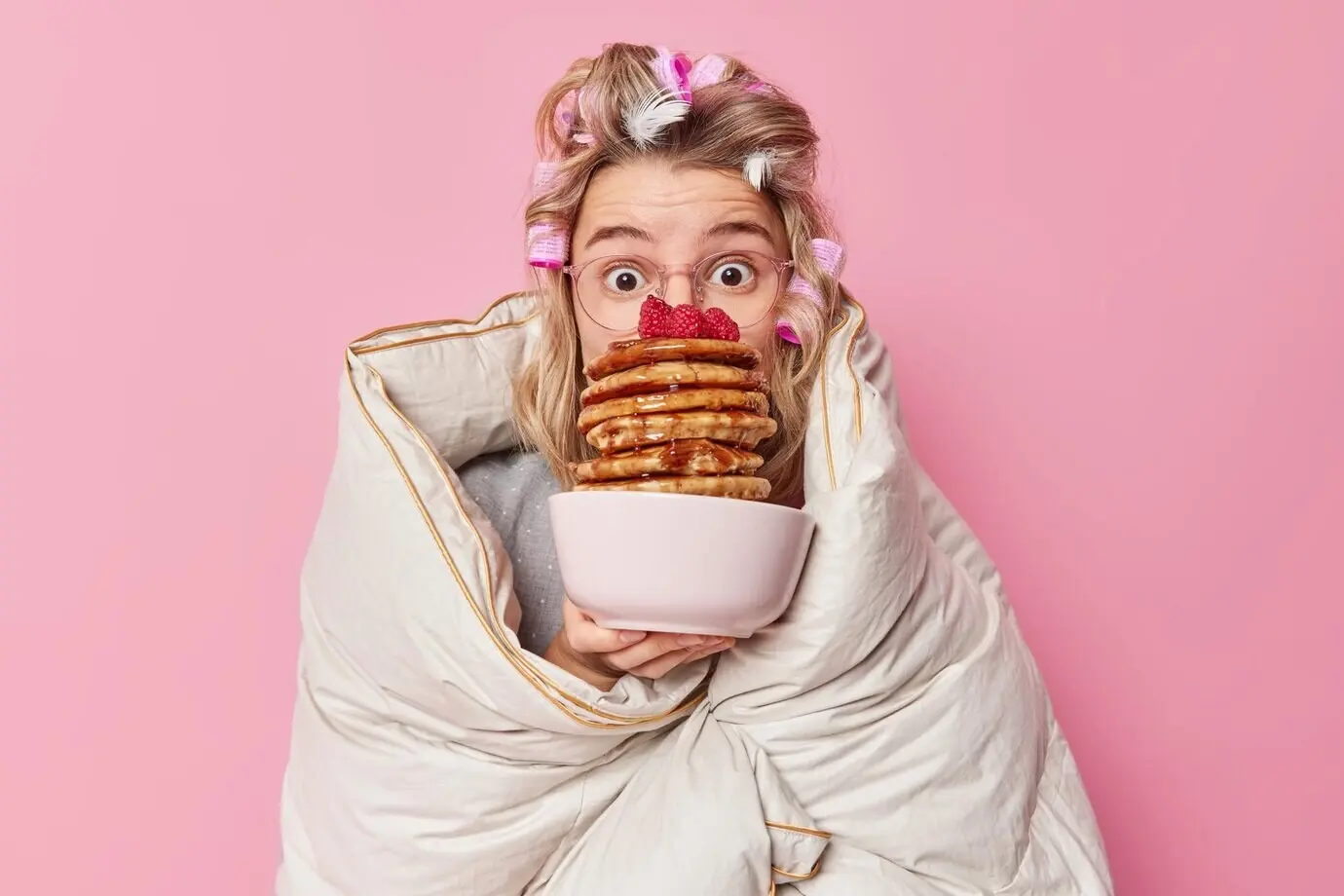 Impressed young European woman with hair rollers, wrapped in a duvet and wearing big spectacles, holds a bowl of appetizing pancakes with syrup, isolated against a pink background. Breakfast and morning time concept.