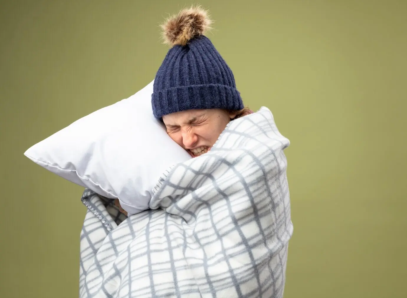 An aching young ill girl wearing a white robe and a winter hat with a scarf, wrapped in plaid, hugging a pillow.